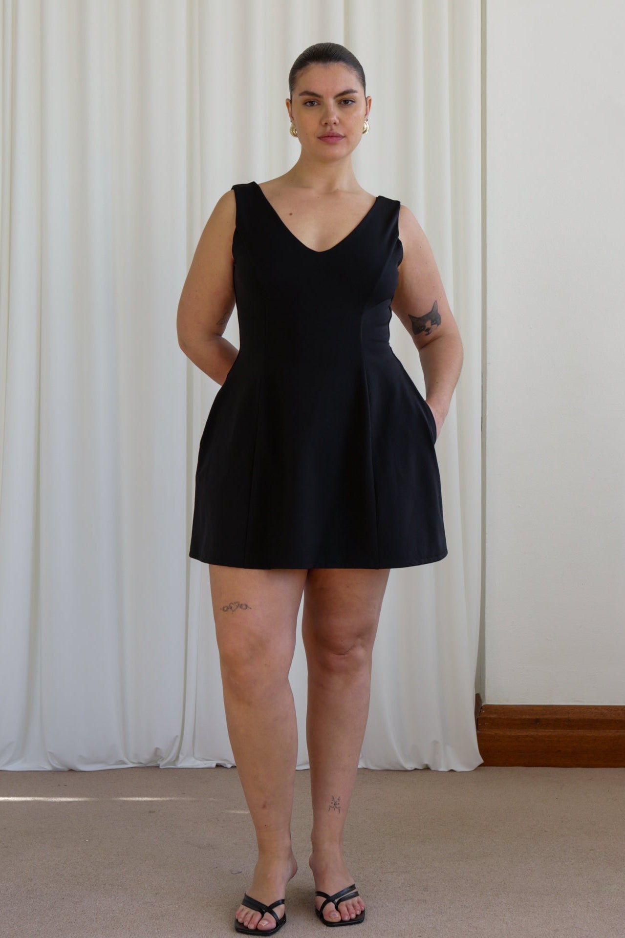A woman standing in a room with white curtains wearing a black mini dress with a V-neckline and no visible sleeves. The dress has a tulip-shaped silhouette and she has hidden pockets on the sides.