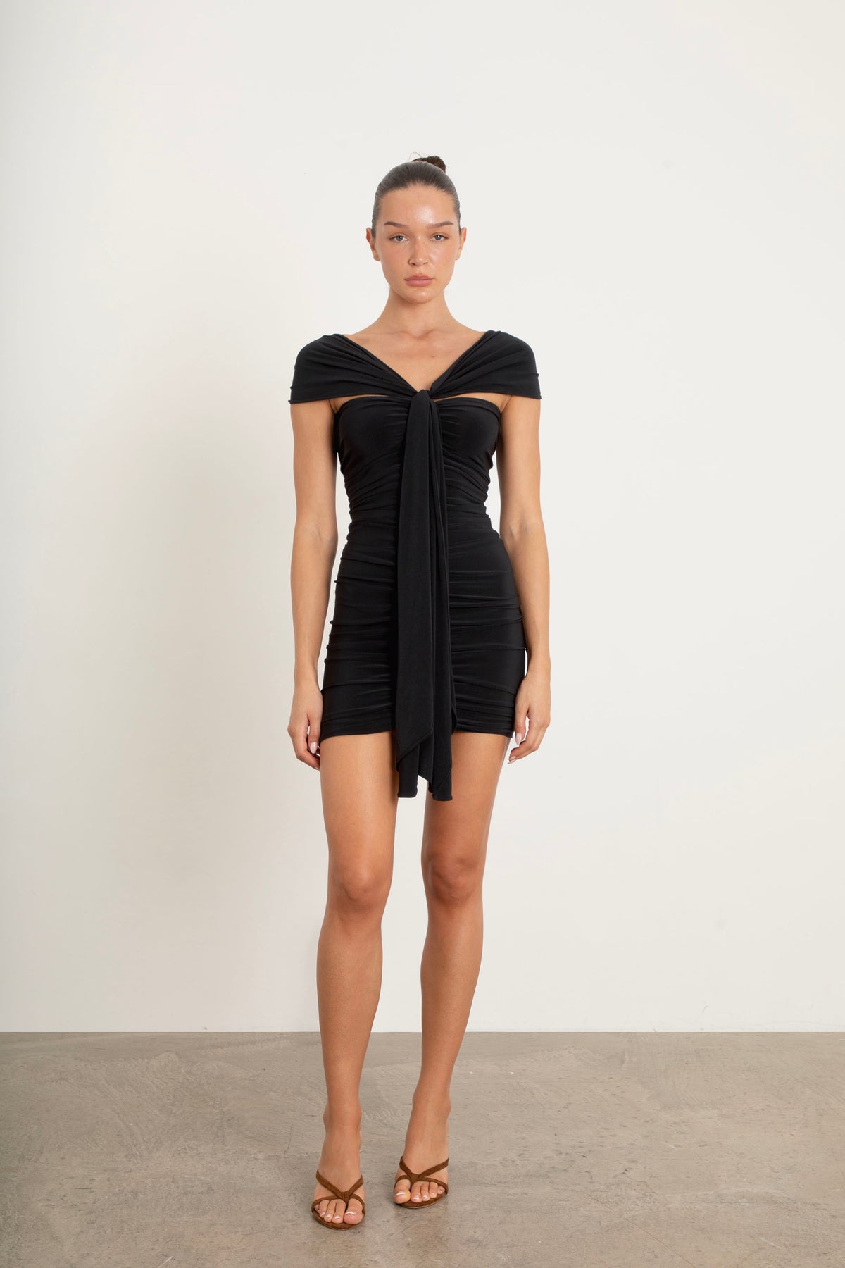 A model wearing a black off-the-shoulder ruched mini dress with a front tie detail, paired with brown strappy heels, standing against a plain off-white wall and concrete floor.