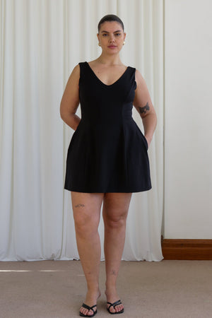 A woman standing in a room with white curtains wearing a black mini dress with a V-neckline and no visible sleeves. The dress has a tulip-shaped silhouette and she has hidden pockets on the sides.